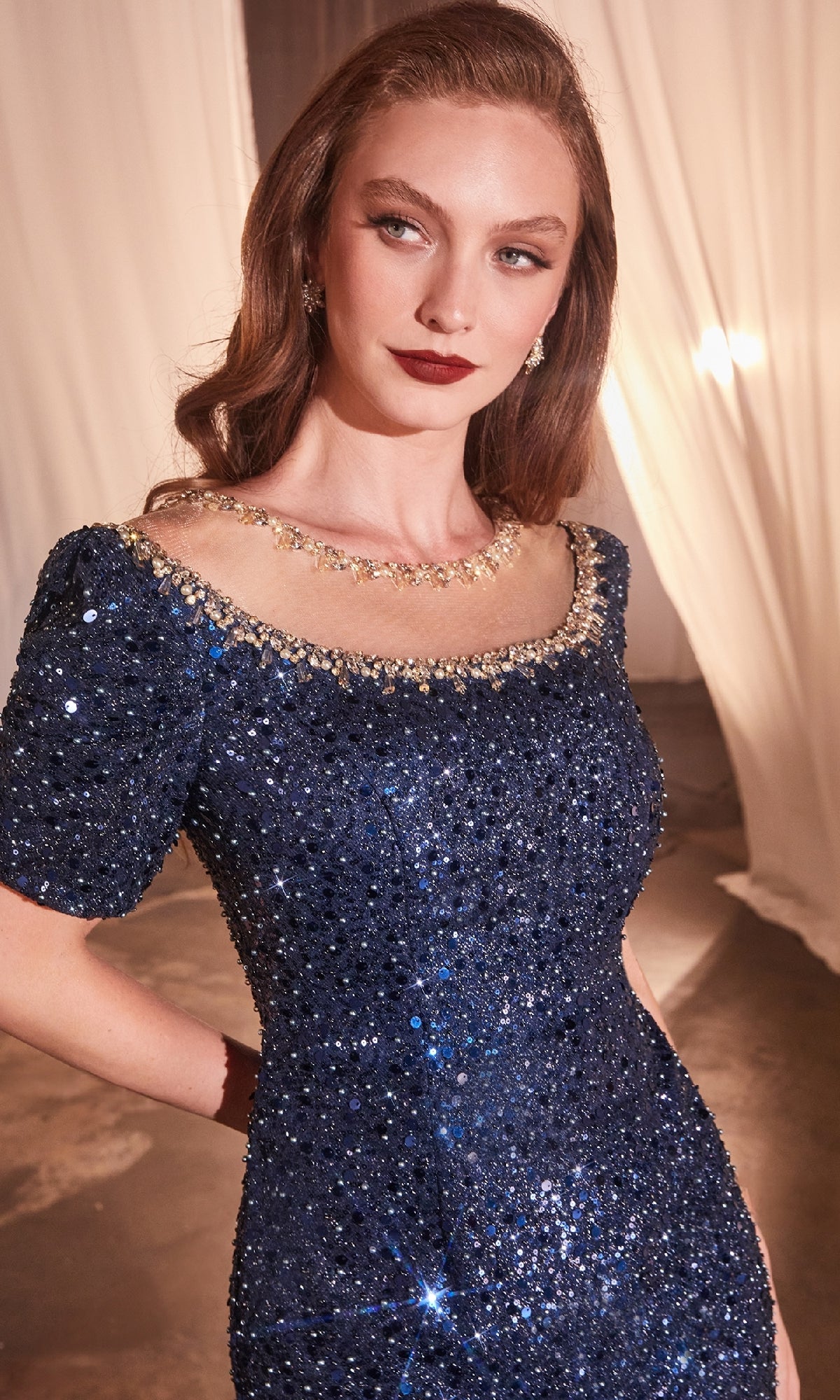 A woman wears the Ladivine CB151 short-sleeve sequin formal dress in navy blue, accessorized with a gold necklace and earrings, standing indoors with draped white fabric in the background.